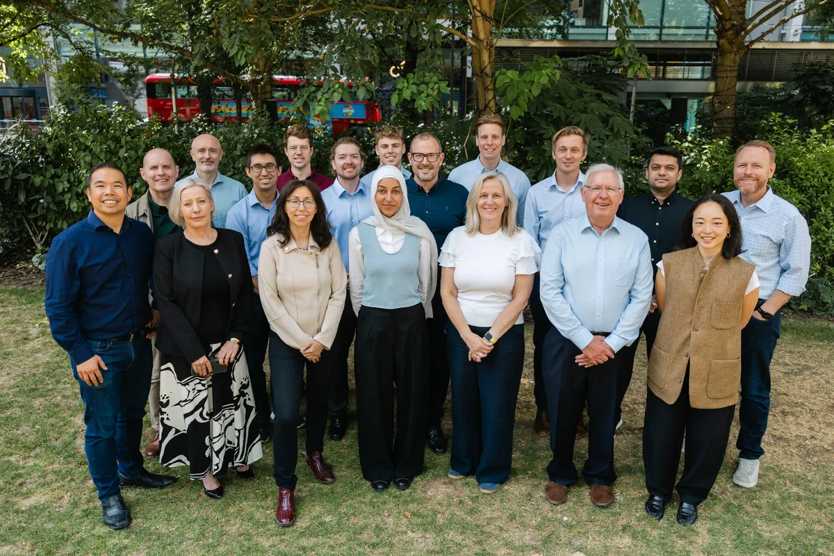 The full Atana team gathered on a lawn under trees, a red London bus passing behind
