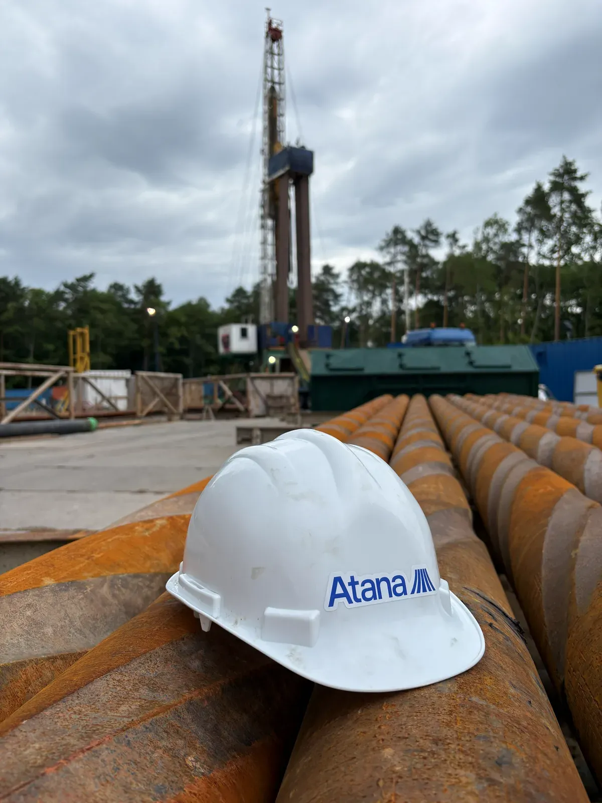 A white Atana hard hat resting on racked, rusted drill pipe, with a drilling rig in the background