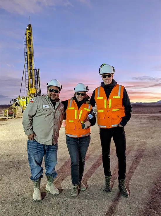Three people in hard hats and hi-vis vests in front of a drilling rig at dusk