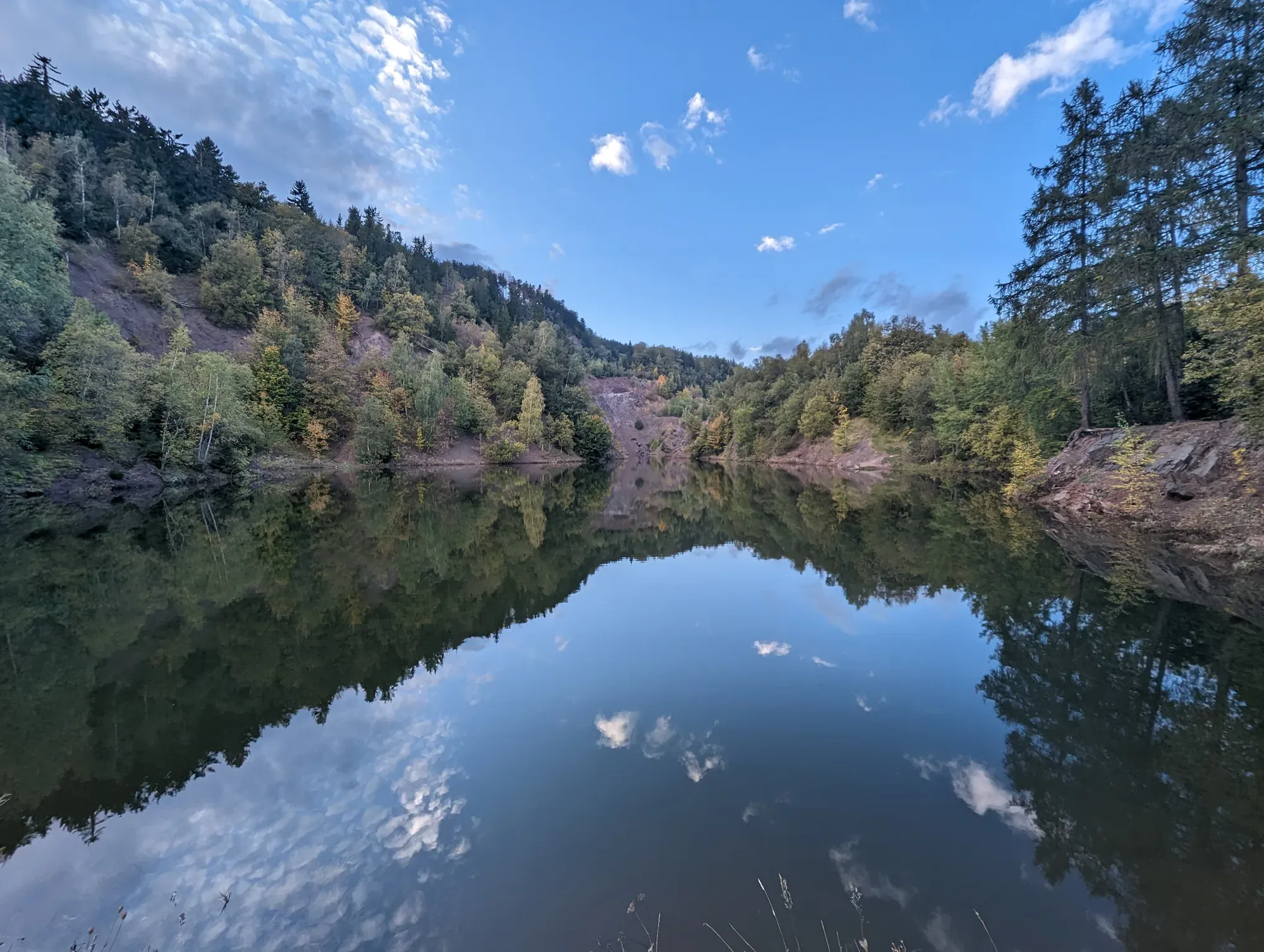 Stiller See zwischen bewaldeten Hängen in frühherbstlichen Farben, mit freiliegendem rötlichem Gestein an den Ufern und dem Himmel, der sich im Wasser spiegelt
