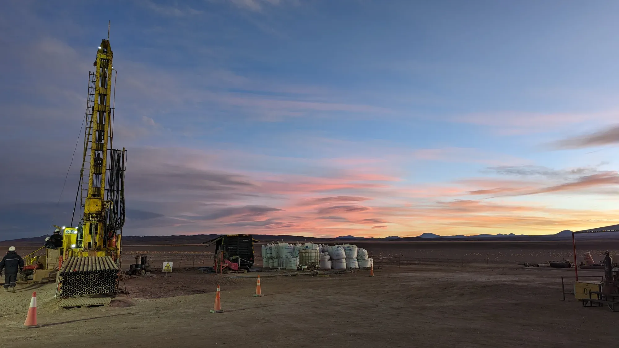 Workers on a yellow drill rig at dusk on a high plain, supplies stacked beside the pad under a pink and orange sunset sky