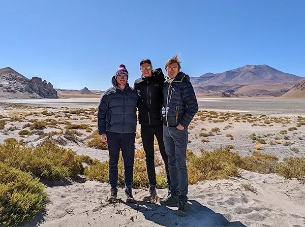 Three team members in jackets on a dry high-altitude plain with mountains behind