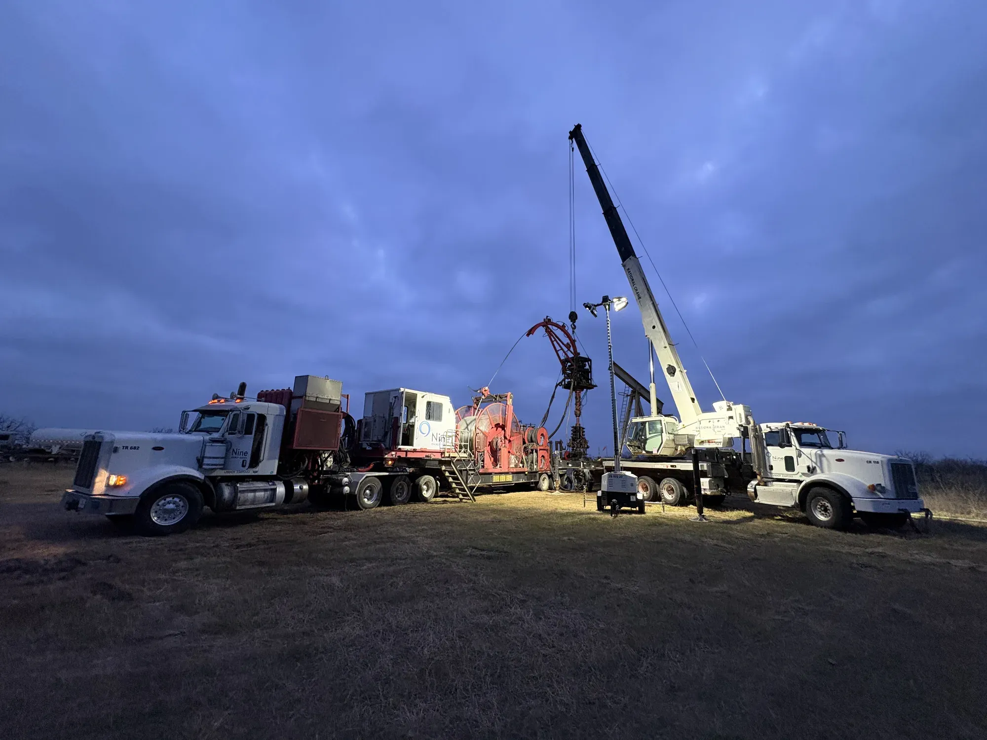 White service trucks and a red coiled-tubing unit floodlit at dusk, a crane boom raised over the wellhead between them